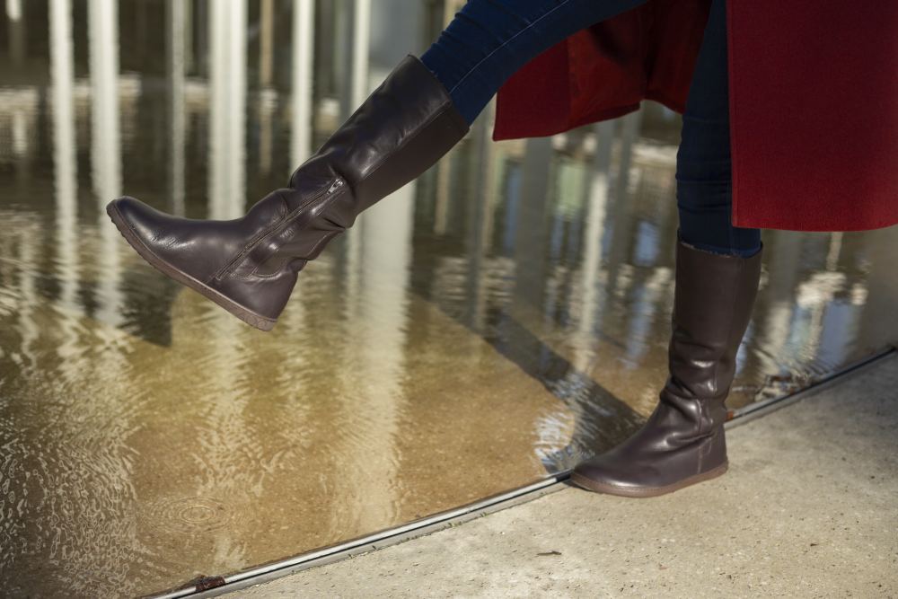 A person wearing dark jeans and a pair of QATCH Winter Brown boots ZAQQ climbs into a shallow pool of water, partially covered by a long red coat. The reflection of the surrounding structures in the water gives the scene an artistic touch.
