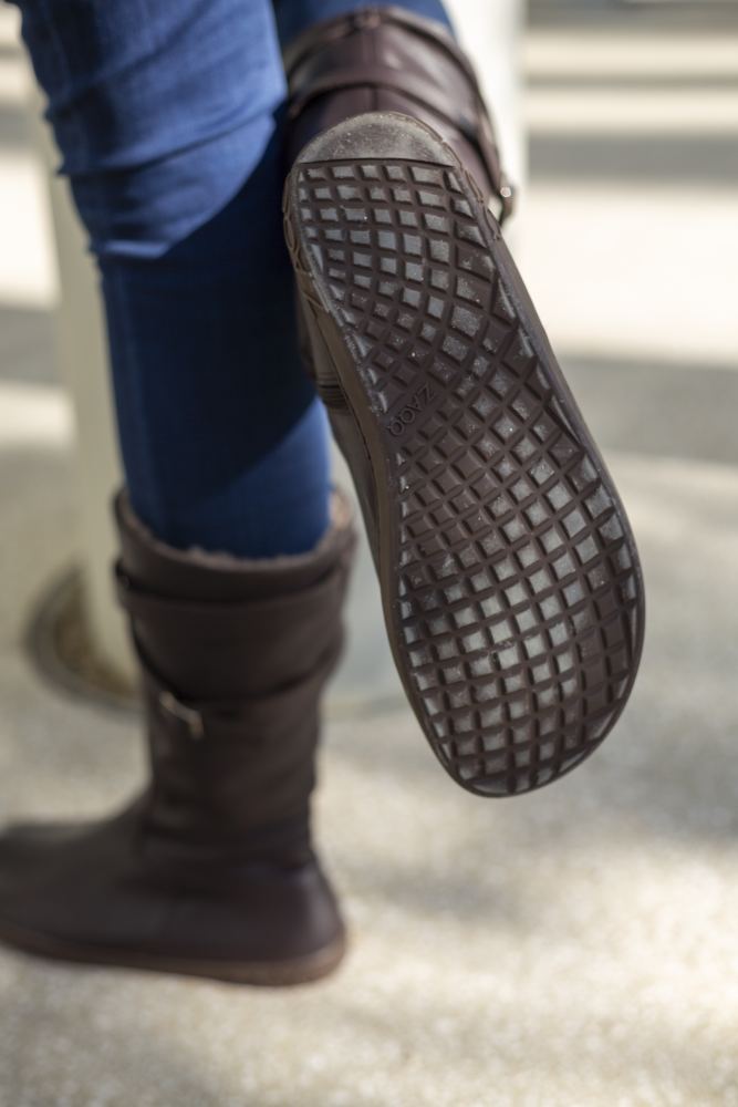 A person wearing the ATTRAQT Winter Brown Boots from ZAQQ and blue jeans, lifts your left foot and reveals the structured sole, which promises excellent wearing comfort. The background is a light surface.
