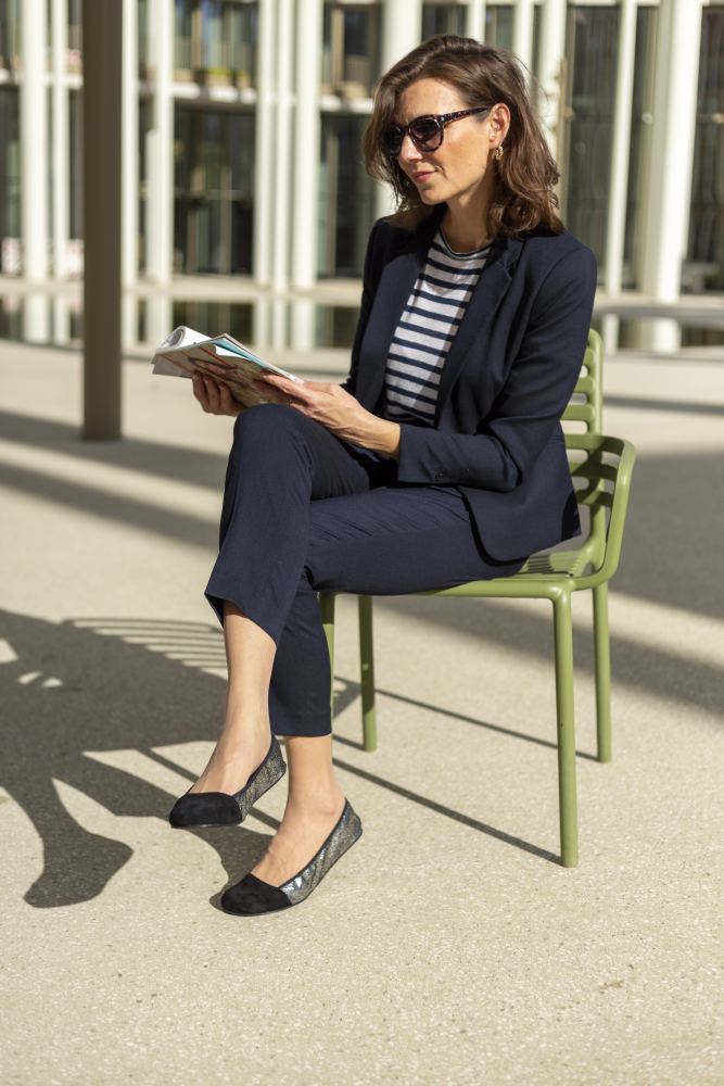 Eine Frau mit Sonnenbrille und marineblauem Blazer sitzt auf einem grünen Stuhl und liest ein Buch. Sie ist an einem sonnigen Tag mit langen Schatten im Freien. In ihrem entspannten Zustand genießt sie den Komfort ihrer schwarzen TWIST Derby-Schuhe von ZAQQ, umgeben von der Kulisse moderner Architektur.