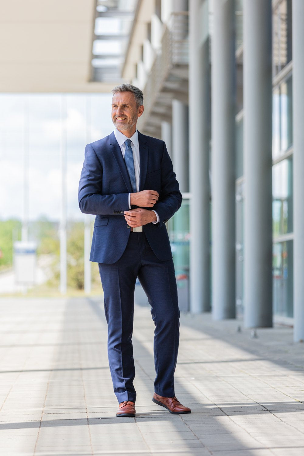 A man in a blue suit and tie stands confidently on a modern, sunlit sidewalk with large stained glass windows and columns. He straightens his sleeve, smiles slightly and wears the comfortable BRIQ Brogue Cognac shoes ZAQQ. The background shows a bright outdoor setting.