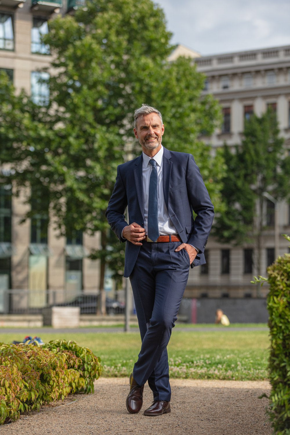 A man in a navy blue suit confidently walks along a gravel path in a green park, with trees and a large building visible in the background. He smiles, his hand in his pocket, and effortlessly combines elegance and casualness. On his feet he wears the elegant DUQE Vegan Antique Brown boots from ZAQQ made of vegan material.