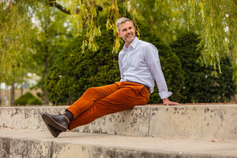 A man with graying hair sits on a low stone wall in a park. He wears a white shirt and orange pants. He wears NEWQUAY Brown shoes ZAQQ, which are made of supple nappa leather. Lush green trees and leaves can be seen in the background, creating a tranquil outdoor backdrop.