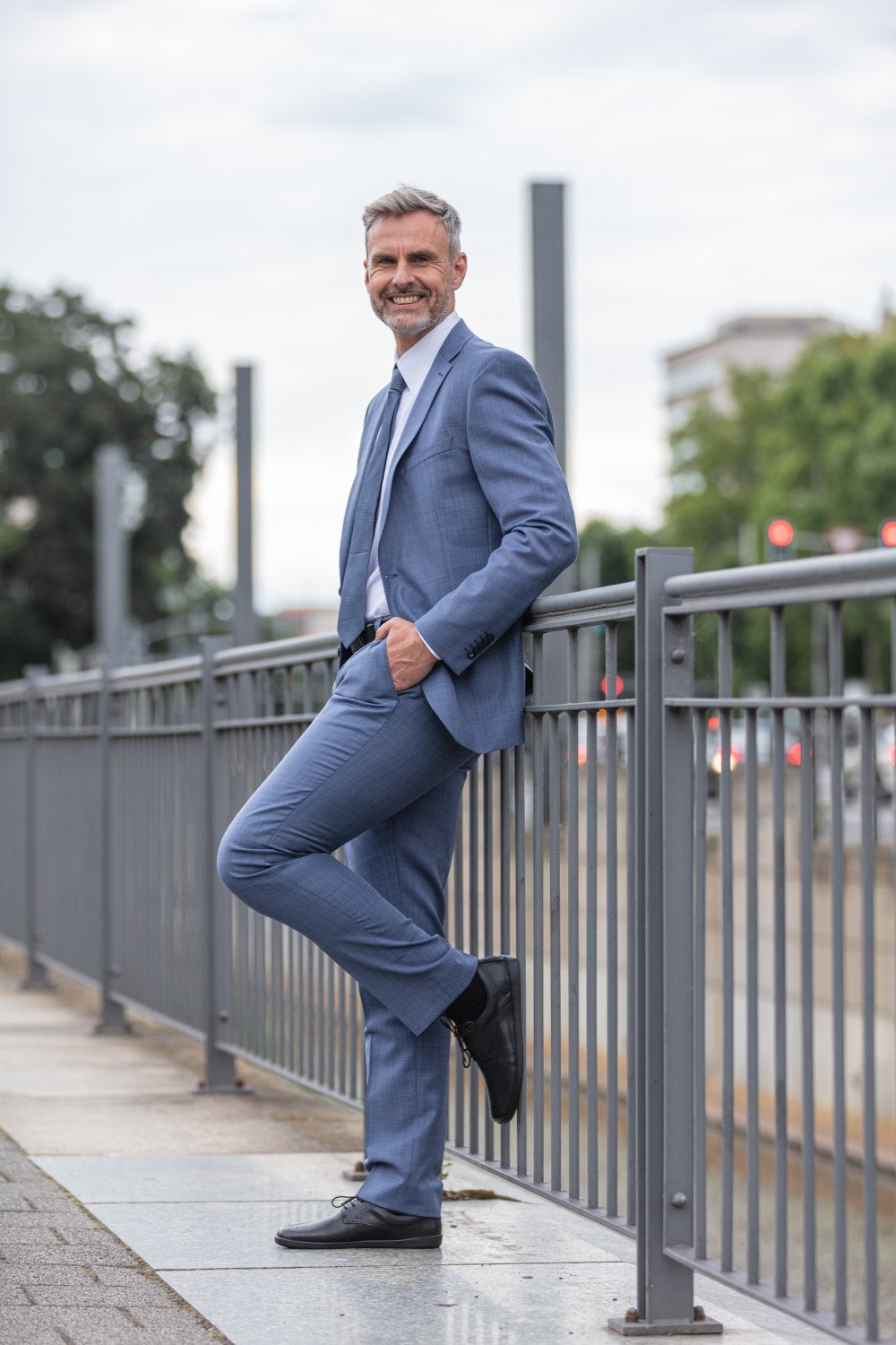 A man in a blue suit leans confidently against a metal railing on a city sidewalk. His black ones ZAQQ PEAQ brogue shoes give it an elegant touch. He smiles while posing cross-legged. Blurred trees, buildings and subtle city traffic can be seen in the background.