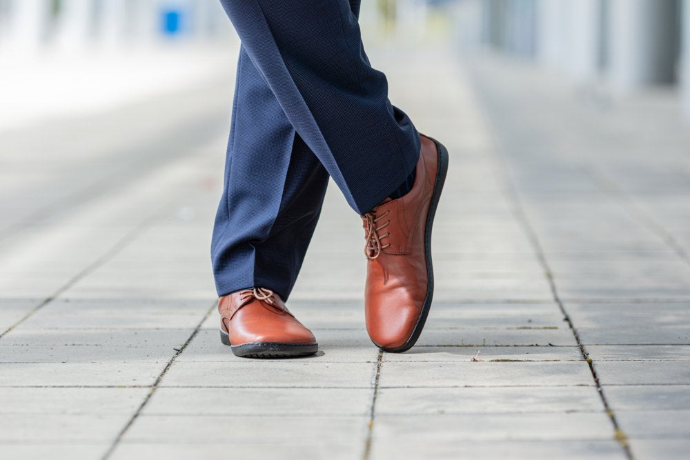 Close-up of a person wearing ZAQQ's PEAQ cognac leather shoes and navy pants walking on a cobbled sidewalk. The image captures a dynamic stride and highlights the style and elegance of these sleek smooth leather shoes.