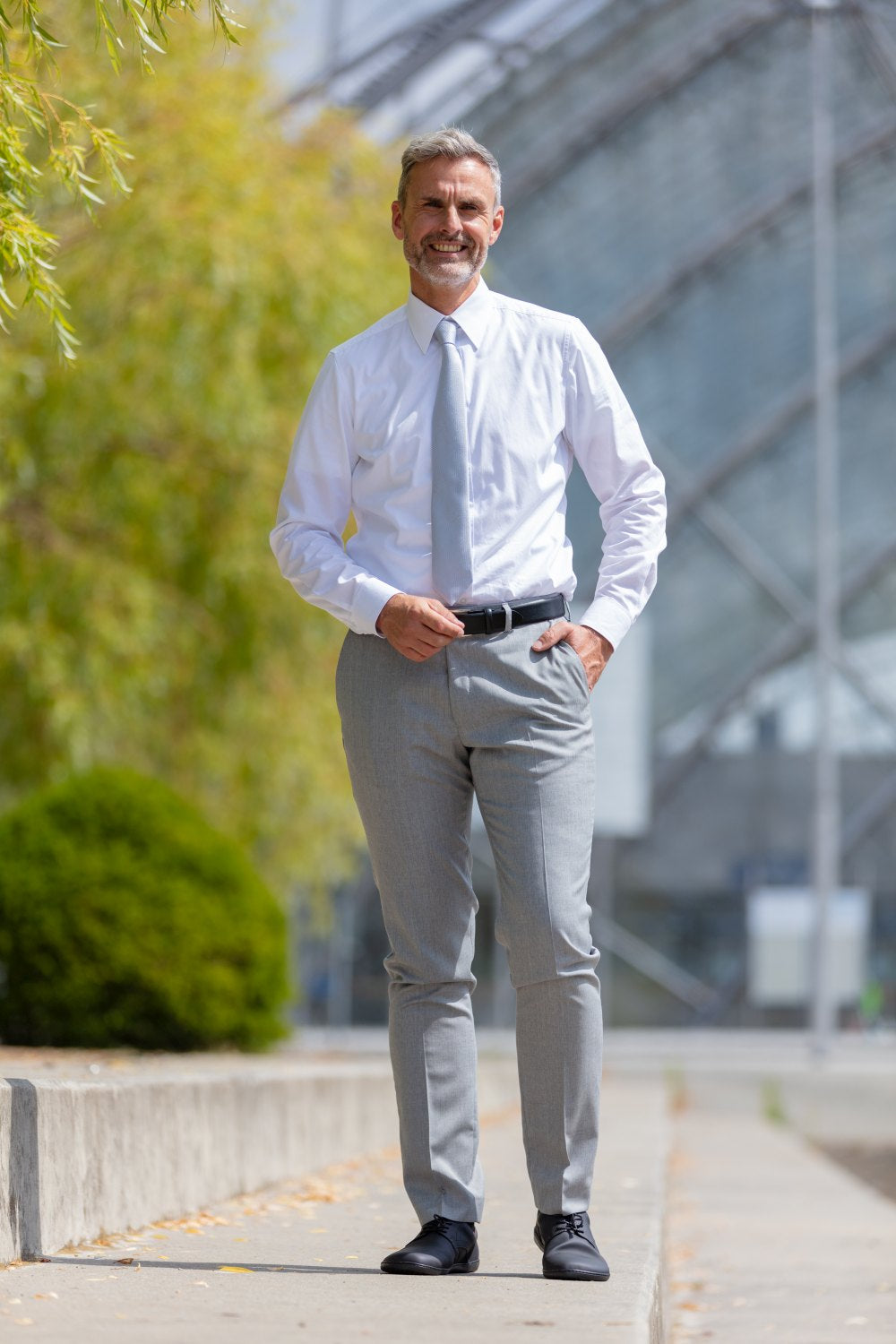 A man with gray hair and a beard stands outside in a modern, urban environment. He wears a white shirt, light gray suit pants and elegant PEAQ Vegan Black shoes ZAQQ. He smiles against a backdrop of glass architecture and greenery. His shoes are equipped with breathable microfiber lining for greater comfort.