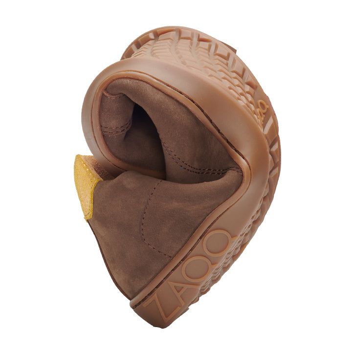 The QOMFY Wide Brown shoe from ZAQQ can be bent in the middle, highlighting its flexibility and superior comfort. This extra-wide model features a chunky sole with a unique tread pattern and embossed lettering on the side. The soft upper is enhanced by a striking yellow label on the inside.