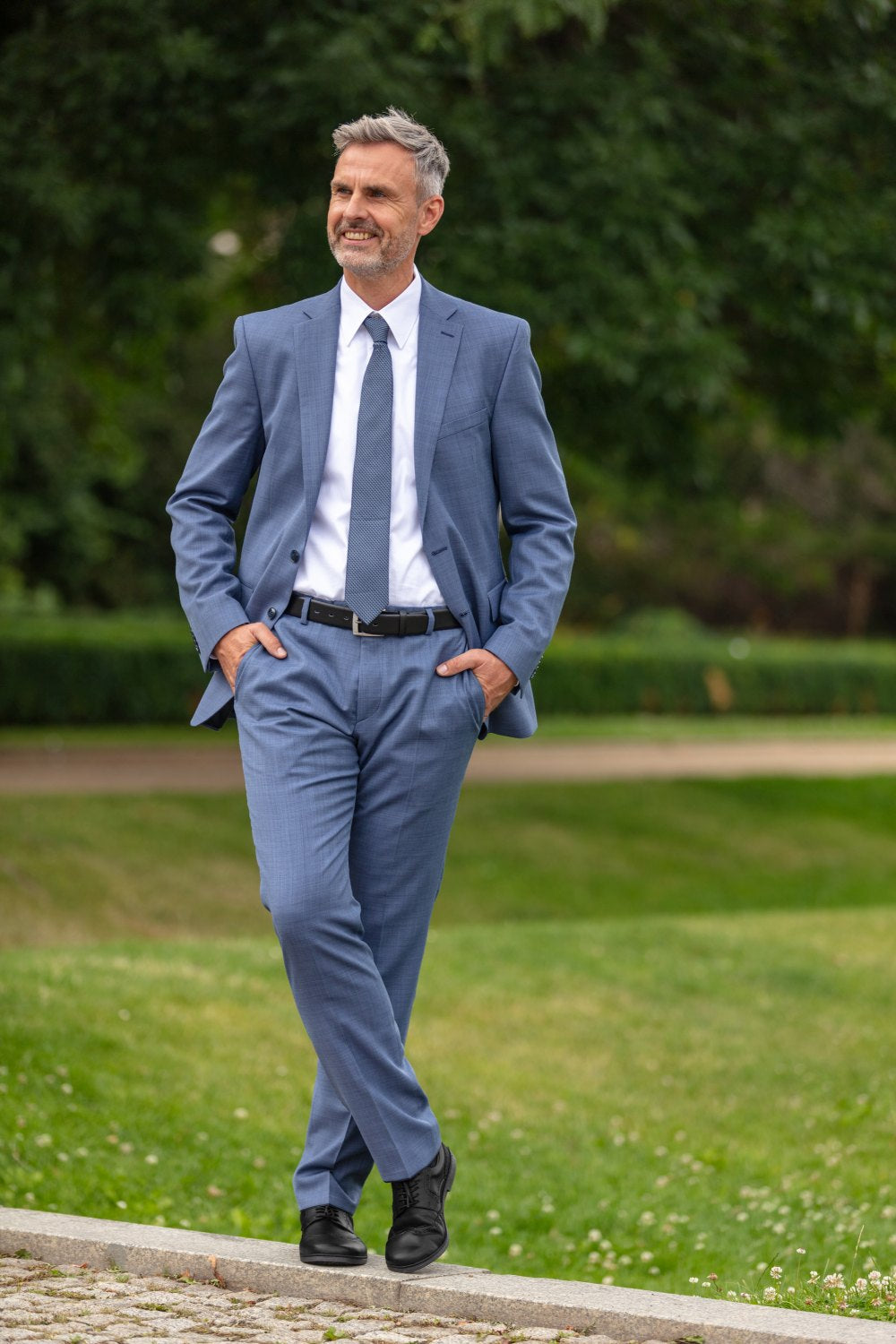 A man in a blue suit and tie walks outside on a path with his hands in his pockets. He is smiling, has gray hair and a beard, and stands against a background of green grass and trees. He wears the BRIQ Brogue Black from ZAQQ, which completes his elegant look.