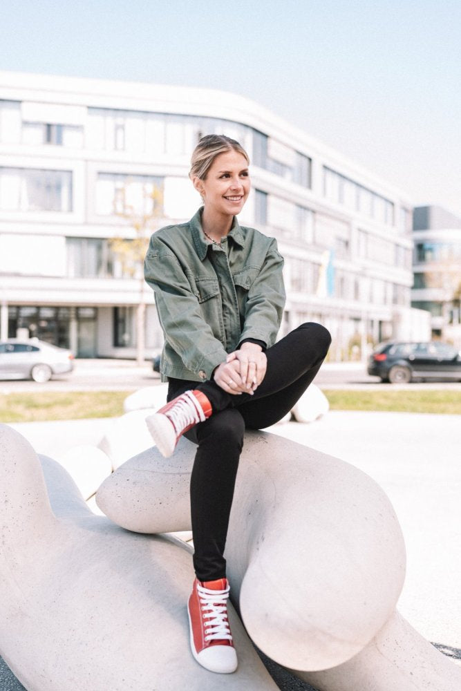 A person with short blonde hair sits on a large, abstract white sculpture outdoors. She wears a green jacket, black pants and red ones ZAQQ CHUQQS sneakers. A modern building and parked cars can be seen in the background.