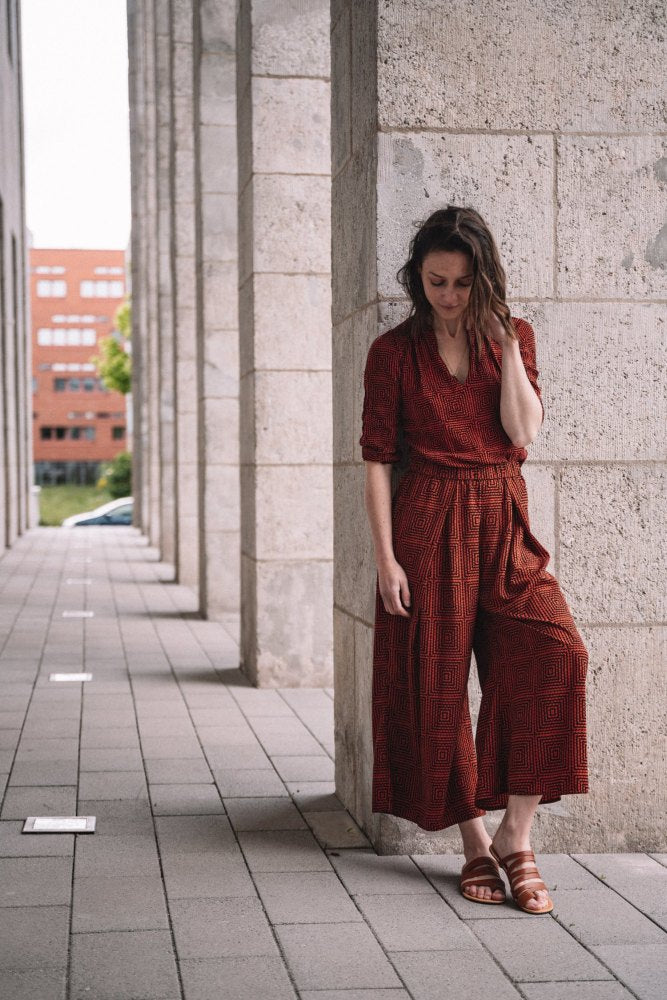 A woman leans against a stone pillar in an outdoor walkway and looks gently downward. She is wearing the FLAQE Brown Jumpsuit from ZAQQ, combined with minimalist sandals. Buildings form a distant backdrop, framing your quiet moment with timeless elegance.