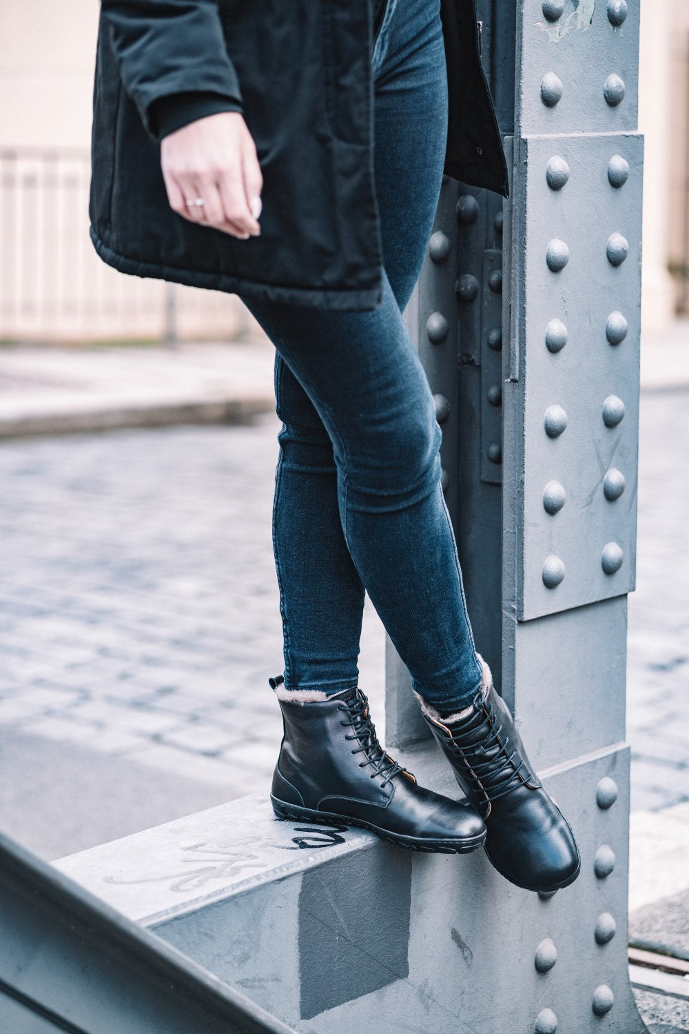 A person in a black coat and skinny jeans leans against a riveted metal beam. She is wearing the QUINTIC Winter Black boots from ZAQQ, which are perfect for the chilly day, and stands on a cobbled city street.