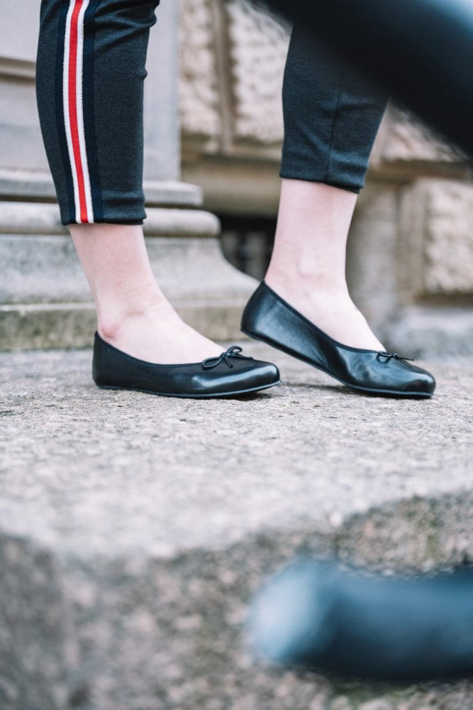A person wearing black ballerina flats from LOOQ ZAQQ and wearing black pants with a red and white side stripe, stands on stone steps partially obscured by a black railing.