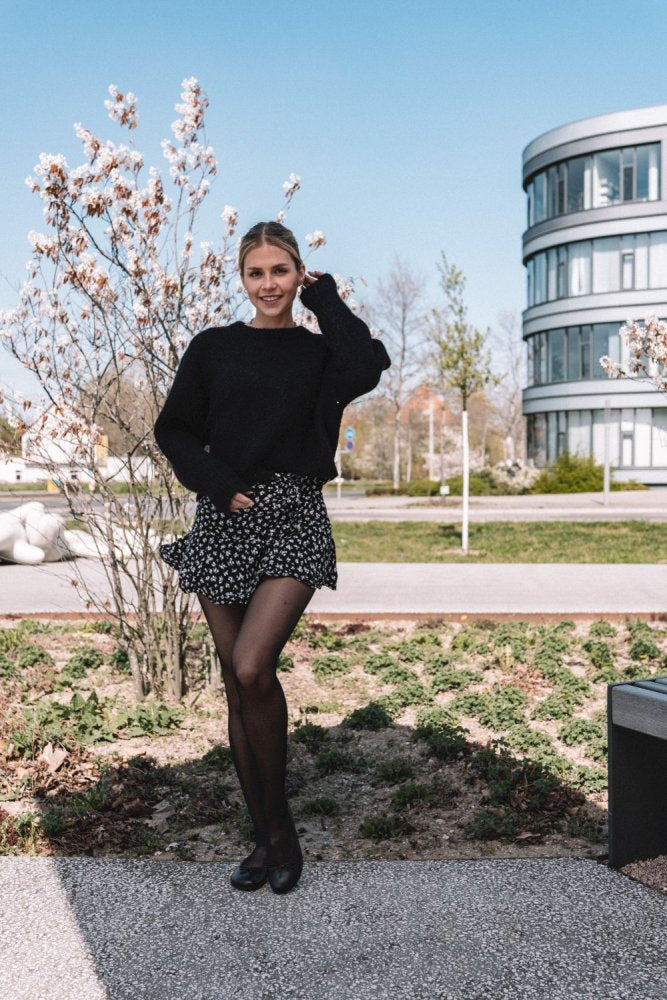 A person stands outdoors smiling, wearing a sweater and a floral skirt ZAQQcollection. The LOOQ Dark Brown outfit adds to her effortless style, reminiscent of a barefoot ballerina. They are framed by flowering trees and a modern building in the background under the clear blue sky.