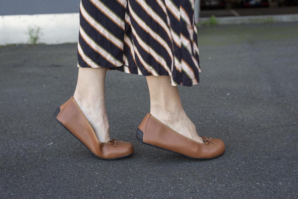 One person wears the LOOQ Vegan Cognac ballerinas with barefoot soles from ZAQQ and strides gracefully across a gray asphalt surface. She also wears a long skirt with black and beige diagonal stripes that embodies style and sustainability.