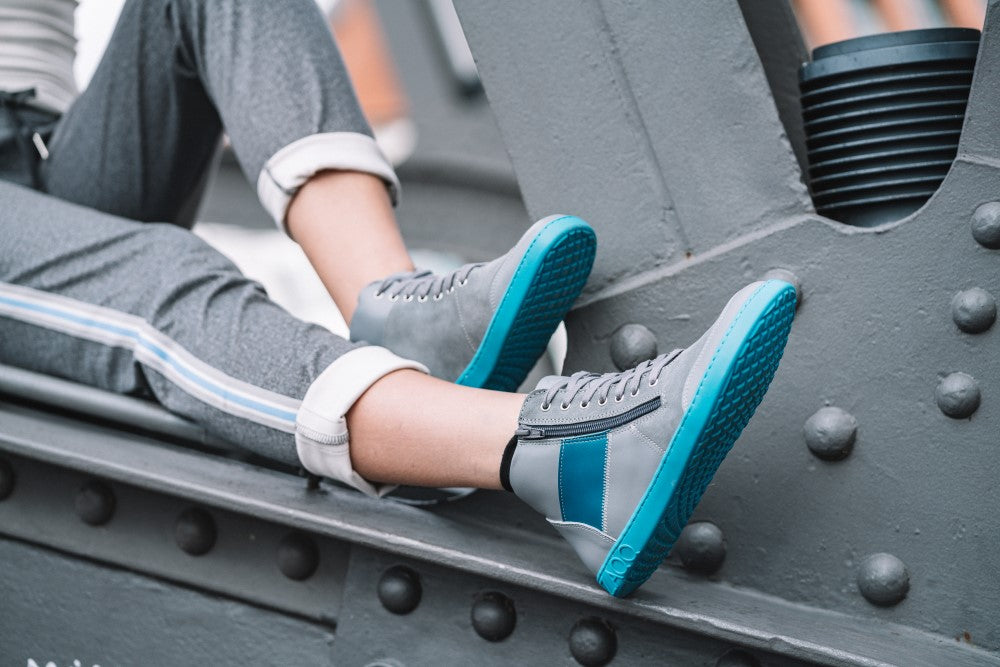 A person dressed in gray sweatpants with the trouser legs rolled up and stylish ZAQQ LUQQ Turquoise sneakers with a blue sole and accents, sit on a metal construction that is reminiscent of a bridge or an industrial site and conveys a relaxed, urban atmosphere.