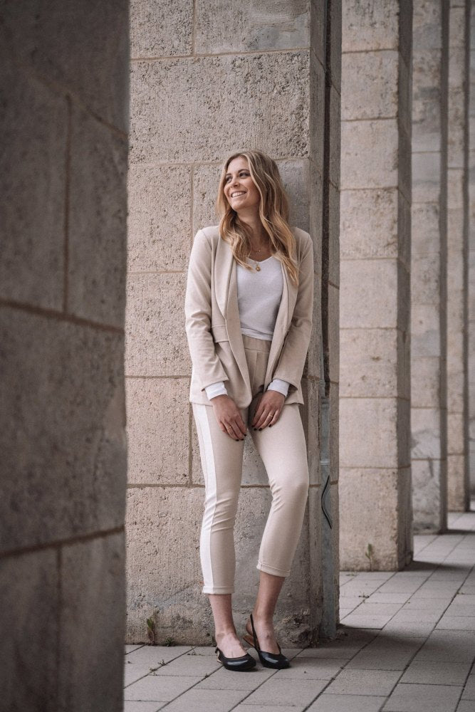 A woman in a light suit stands smiling in front of a stone wall, her appearance as graceful as that of a ballerina. She's standing outside on a cobbled square with tall pillars, feeling relaxed and happy in her stylish PINQ Black shoes ZAQQ.