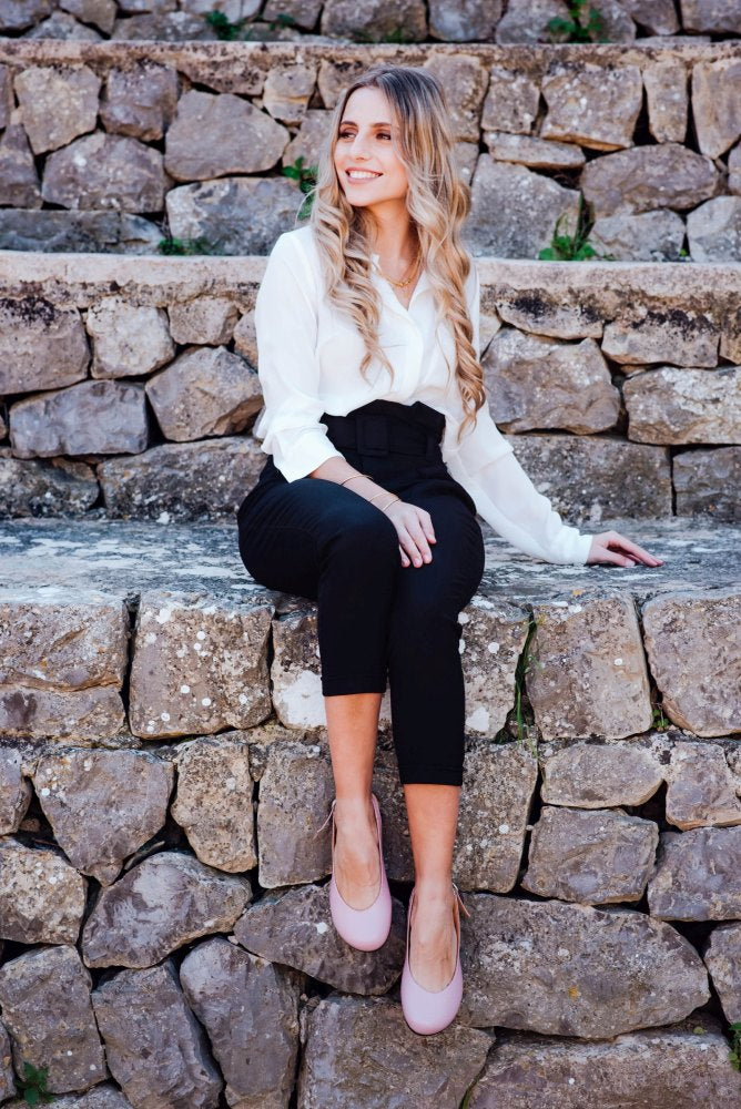 A woman with long, wavy hair sits on a stone wall. She is wearing a white blouse, black pants and feminine PINQ Candy sandals from ZAQQ. She smiles and looks to the side, with terraced stones behind her.