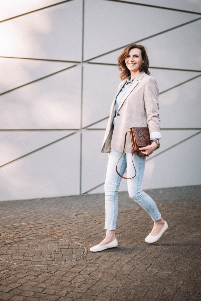 A woman in a light check blazer and jeans strolls confidently in her beige ones ZAQQ QARMA shoes over a cobblestone path. She holds a brown handbag and smiles in front of the modern geometric wall, her barefoot shoe treading lightly on the sunlit stones.