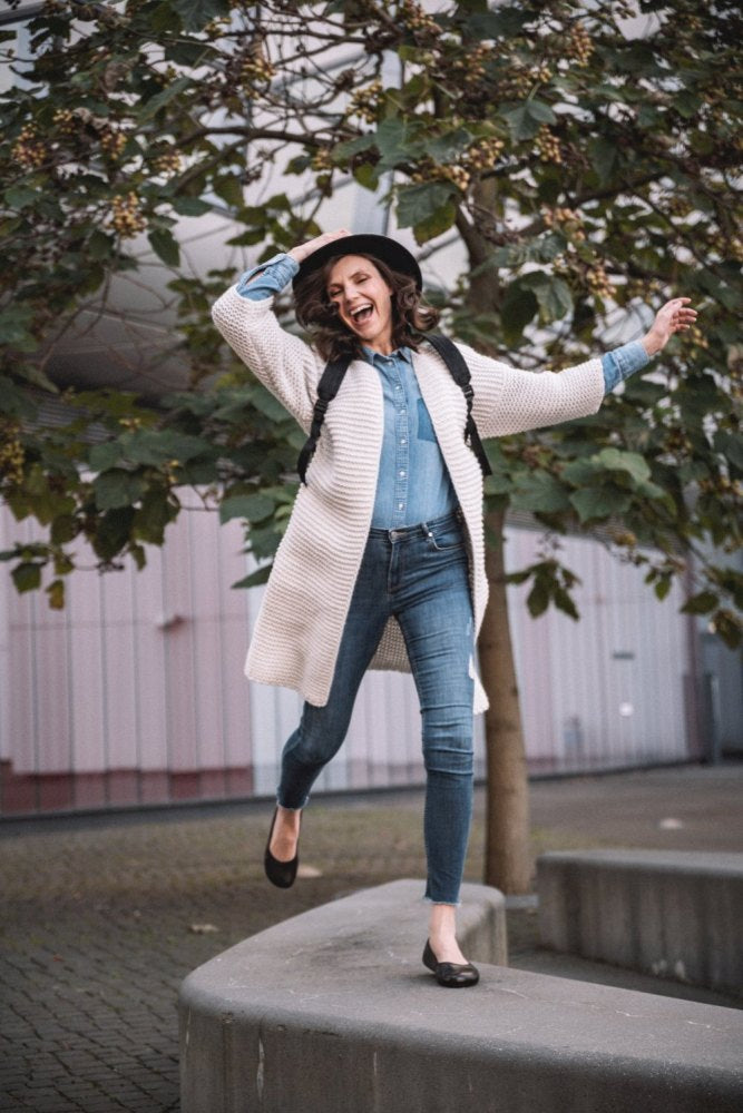 A cheerful person in a hat and long cardigan walks energetically along a curved low wall surrounded by trees with green leaves. She is wearing a jeans outfit and dark brown QARMA shoes ZAQQ, which are reminiscent of barefoot ballerinas. She is carrying a backpack. The exterior facade of the building provides a fascinating backdrop.