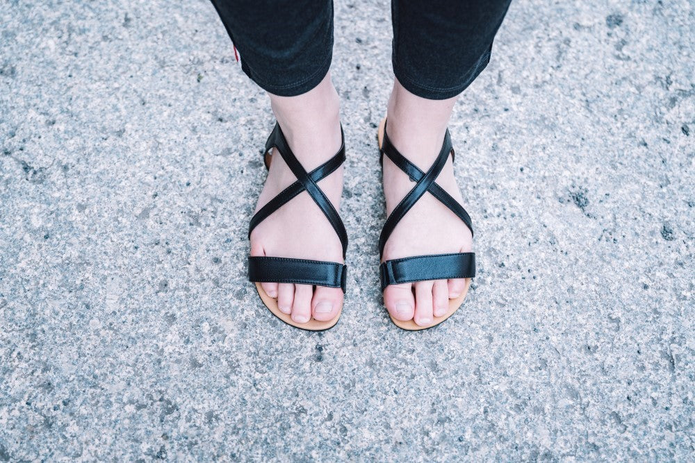 A person who wears the QEE-Black strappy sandals from ZAQQ wearing, stands on a textured, light gray sidewalk. The stylish QEE Black sandals have multiple criss-cross straps over the feet and go perfectly with dark trousers.