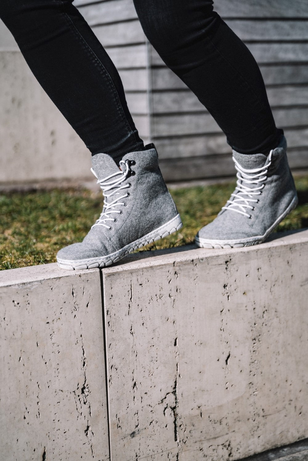 A person wears gray QICE high-top sneakers ZAQQ with an outdoor sole and pairs it with black pants as she balances outdoors on the edge of a concrete ledge where a hint of green grass can be seen in the background.