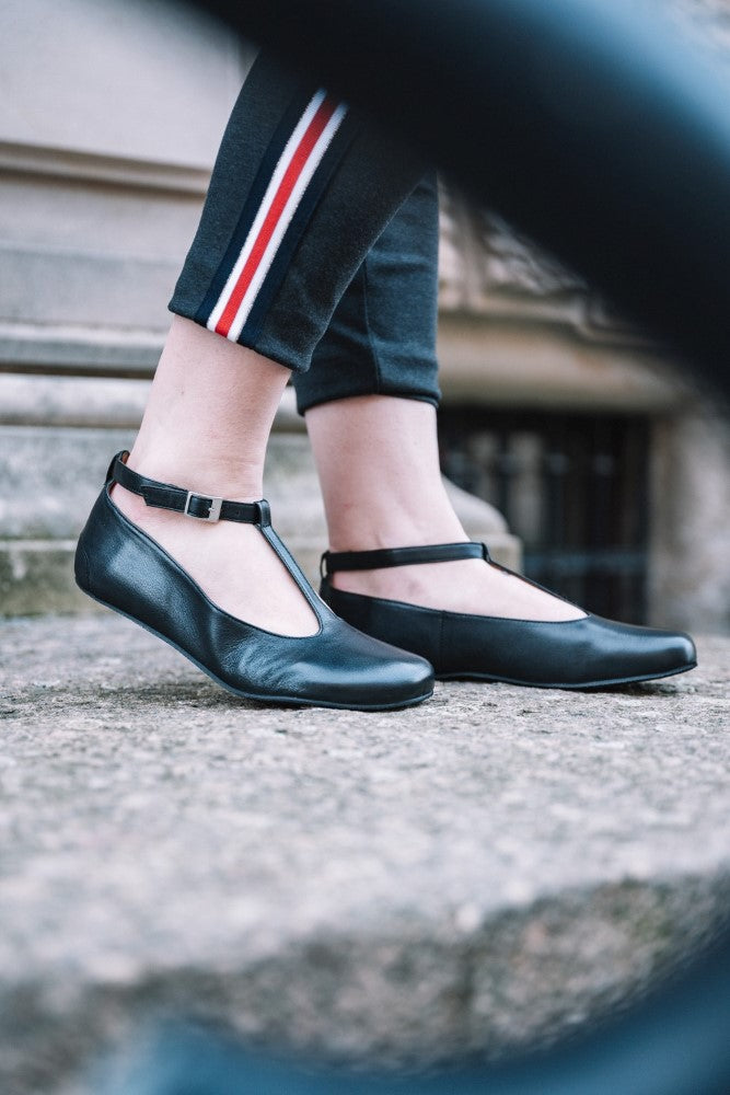 A person wearing QITE Black shoes ZAQQ with an ergonomically shaped footbed stands on a stone surface. She wears black pants with red and white stripes. The image is framed by a blurred object in the foreground.