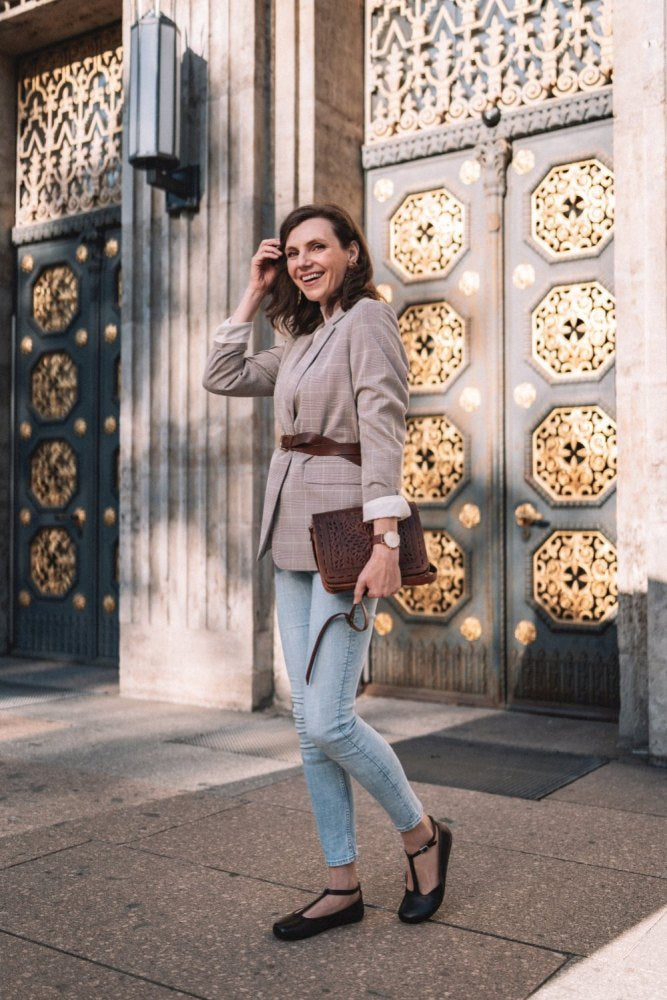 A person in a checkered blazer and jeans stands on the street and holds a structured clutch. In the background you can see doors with an elaborate gold pattern. She wears a watch on her wrist and stylish black shoes made of smooth leather ZAQQ, known as the QITE Dark Brown model.
