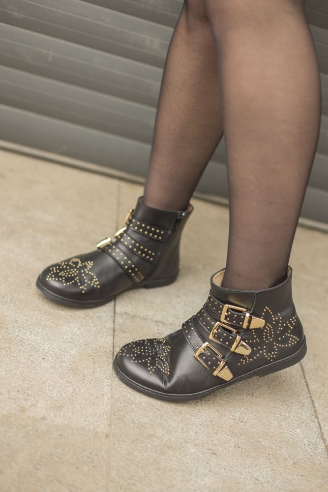 One person wears sheer black tights and ZAQQ's QLANCE Black Gold barefoot boots, decorated with gold studs and straps. The boots feature intricate patterns and are fastened with three gold buckles on each side. The concrete floor and the metal door in the background perfectly highlight the black and gold accents of the ensemble.
