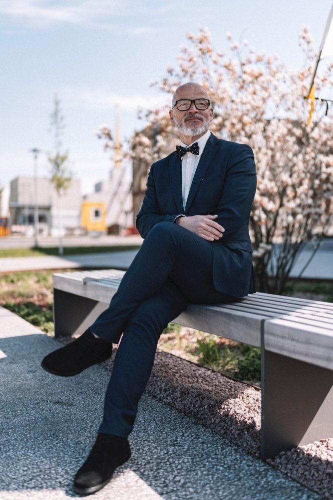 A man in a suit, glasses and a bow tie sits on a bench. He wears stylish black QLASSY shoes from ZAQQ, Crosses your legs and appears relaxed. In the background, flowering trees and a clear sky decorate the sunny day.