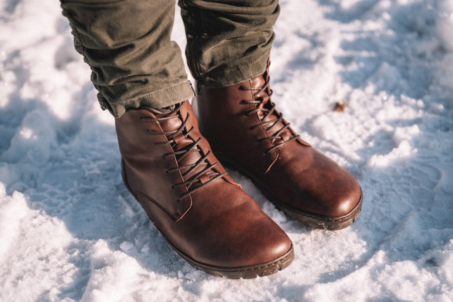 A close-up shows a person wearing QUINTIC Winter Vegan Groove Leather Boots ZAQQ wearing olive green pants and standing on snowy terrain. The vegan lace-up shoe features a durable sole designed for optimal performance in winter conditions.