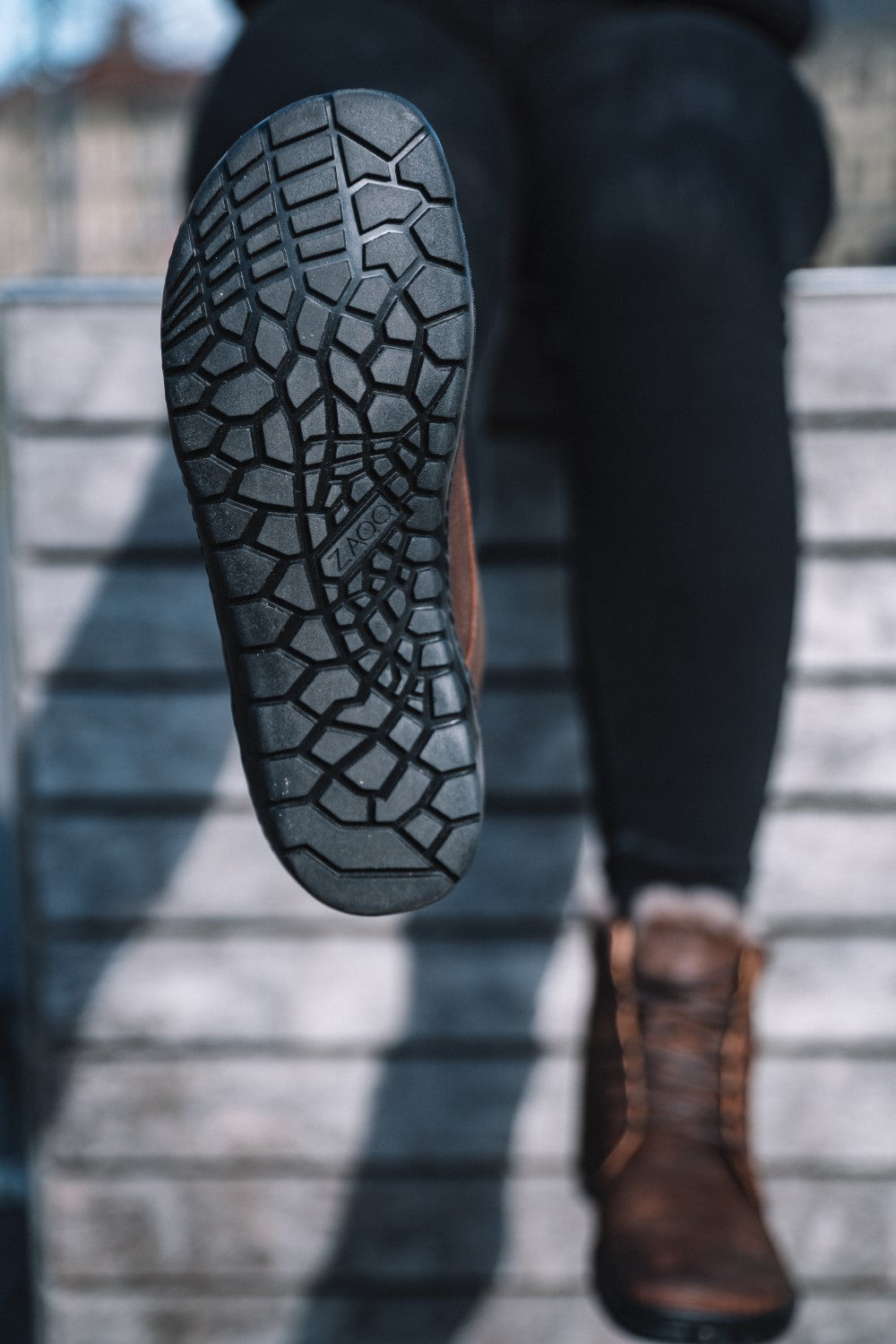Close-up of the sole of a raised ZAQQ QUINTIC Winter Velor Brown waterproof boots with detailed tread pattern. The sheepskin-lined boot is worn by someone sitting on a wooden bench in dark trousers. The hexagonal design on the sole stands out clearly against the slightly blurred background.