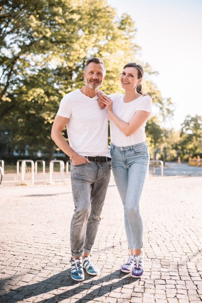 A couple stands outdoors smiling on a sunny day, both wearing white shirts and jeans. The man who made RIQ Purple Sneakers from ZAQQ peeking out, has his hands in his pockets. The woman puts her hand on his shoulder. Trees and a clear sky form the backdrop of this casual moment.