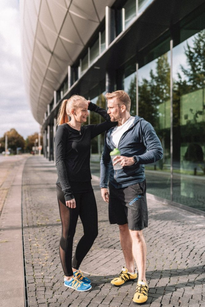 A man and a woman in sportswear stand in front of a modern building with large glass windows. The man is holding a cup and they appear to be deep in conversation. Both wear the RIQ Yellow running shoes ZAQQ, which are known for their flexible feel on the cobbled sidewalk.