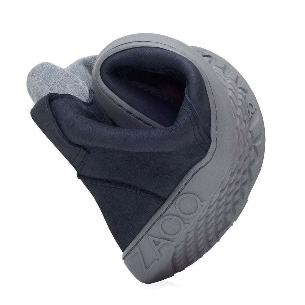 A flexible black ROQA cloud sneaker from ZAQQ with a gray sole that curves in the middle to highlight its comfort. The textured pattern on the sole includes the word printed on the side ZAQQ, all against a pristine white background.
