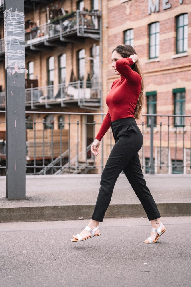 A woman walks confidently down a street. She is wearing a red long-sleeved shirt and black pants, with minimalist ones ZAQQ SLIQ Silver Sandals. With her long hair flowing, she strolls past a railing; brick buildings can be seen in the background.