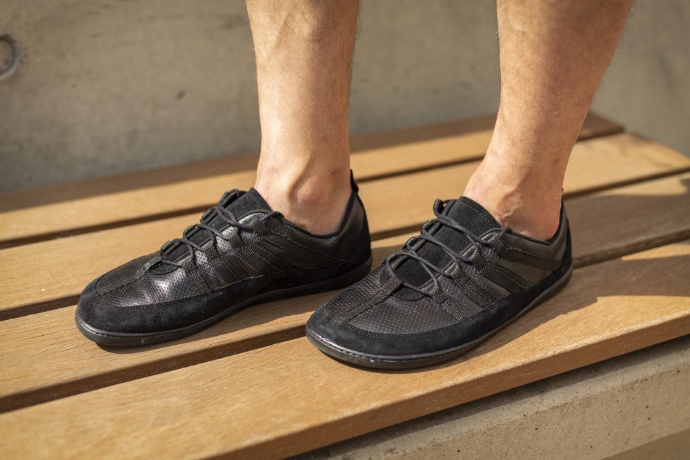 A person stands on a wooden bench and wears the SPARQ Low Black sneakers from ZAQQ. These stylish trainers feature an elegant lace-up design with a combination of mesh and leather, exposed stitching and a minimalist style. The person's bare legs suggest a casual environment.