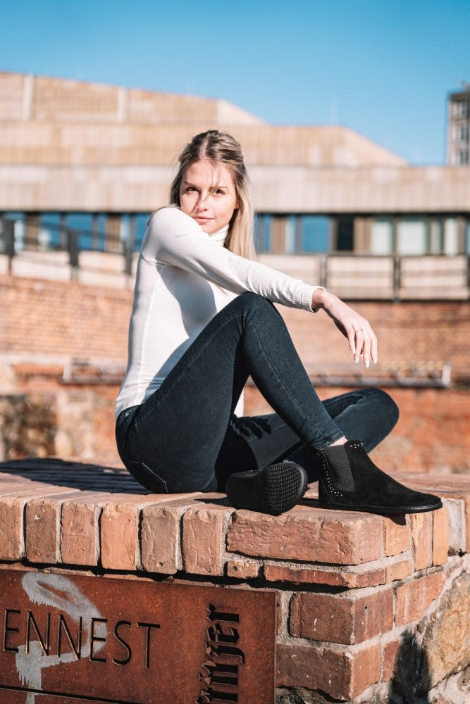 A woman in a white long-sleeved shirt and dark jeans sits on a brick wall and wears ZAQQ's SPARQLE Shine Velor Black Ankle Boots, which are decorated with subtle gemstones. She stands outdoors against a blurred urban background under a clear blue sky.