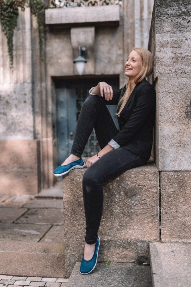 A person with long hair dressed in a black outfit and blue SUQAR Mare shoes ZAQQ, sits on a stone ledge in front of an old building. She smiles and looks forward, one foot resting on the ledge and one hand on her knee.