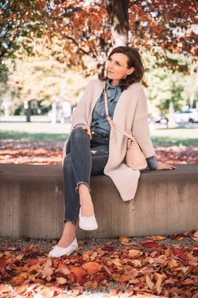 In an autumn setting, a woman wearing a denim shirt, beige cardigan and ripped jeans sits on a low wall. Her light pink shoulder bag pairs well with her white SUQAR sneaker-soled sneakers from the brand ZAQQ. Colorful foliage covers the ground and in the background are trees with orange leaves.