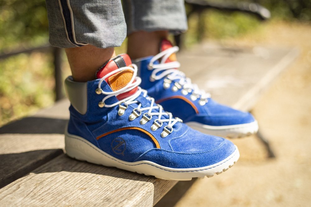 A close-up of a person wearing ZAQQ's TERRAQ Azure waterproof high-top sneakers in bright blue tones, with white laces and red accents. These suede shoes are paired with cuffed jeans while the subject stands on a wooden bench in a sunny outdoor setting.