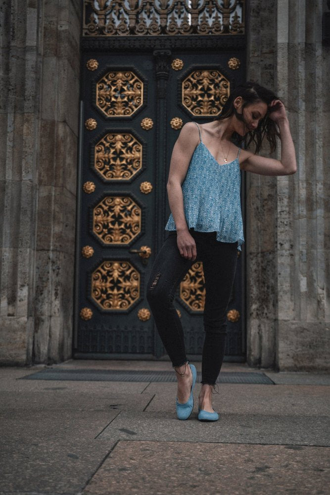 A woman with long hair stands in front of an ornate door with gold details. She is wearing the sleeveless TWIST Light Blue Top from ZAQQ, combined with black jeans and flexible shoes. She looks down and gently touches her hair in a setting that exudes an elegant, atmospheric atmosphere.