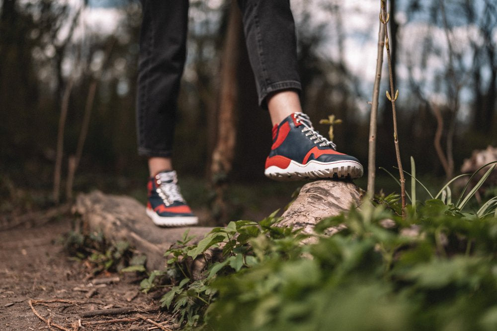 A person in black trousers and the TREQ Red Waterproof Sneakers, which are made as barefoot shoes ZAQQ steps on a fallen tree trunk in the forest. Green plants surround the tree trunk, trees can be seen in the blurred background.