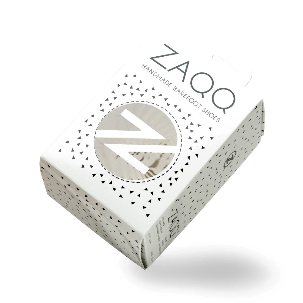 A white shoe box from ZAQQ, Decorated with black triangle patterns, features a round window that showcases the SOQQ Toffee shoes inside. The “Handmade Barefoot Shoe” label highlights the flexibility that goes into each pair.