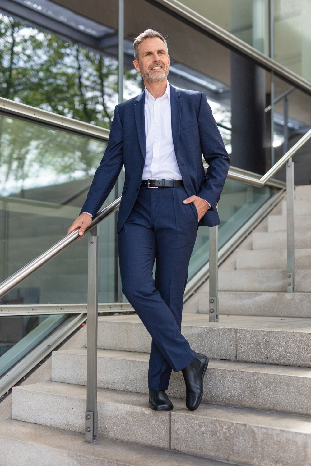 Ein Mann in blauem Anzug, weißem Hemd und schwarzen EQUITY Brogue-Schuhen von ZAQQ steht selbstbewusst auf einer Treppe. Eine Hand in der Tasche und die andere auf dem Geländer, blickt er nach oben und lächelt vor einem Hintergrund aus Glas und Grün.