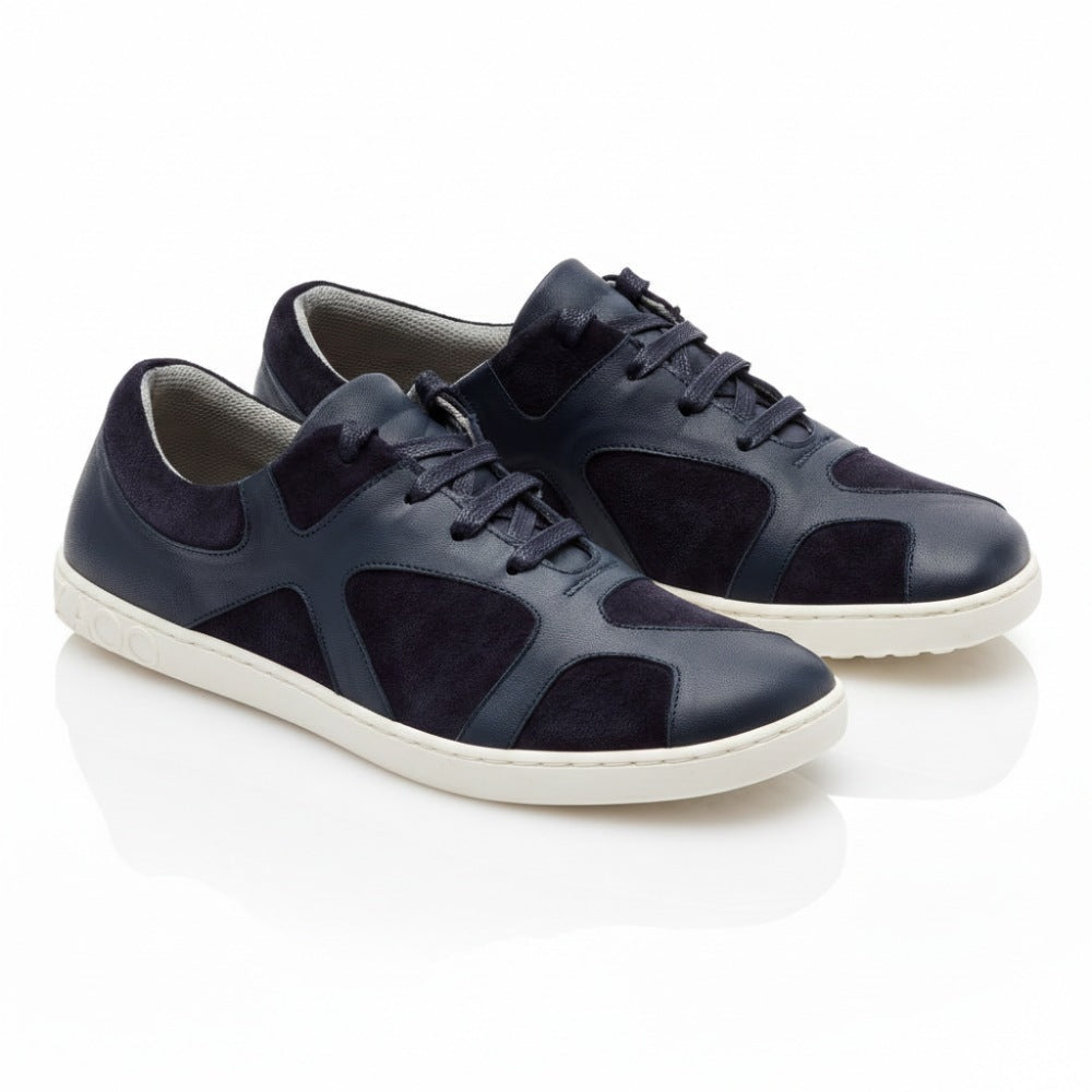 The QAANAAQ Blue sneakers from ZAQQ are made of dark blue leather and suede, have white soles and a lace-up closure in a casual design. They are made of breathable material and stand on a white reflective surface.