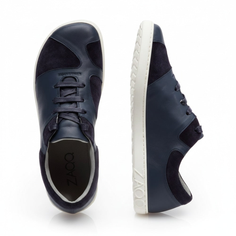 Here the QAANAAQ Blue sneakers from ZAQQ presented. A shoe made of elegant suede stands upright in side view and has a white sole with the ZAQQ-Logo. The other shoe is seen from above, highlighting its lace-up design and simple finish.