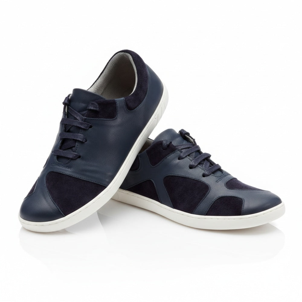 The QAAANAAQ Blue from ZAQQ is a pair of stylish navy blue sneakers with a white sole, made of leather and suede. One shoe is lightly supported on the other to highlight its design and structural details.