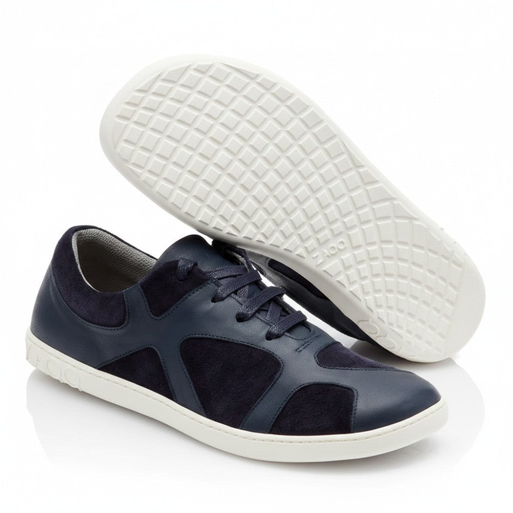 A pair of navy blue sneakers known as QAANAAQ Blue and made of breathable material with gray soles is presented on a white background. One shoe stands upright and shows the side and top views, while the other shows the textured sole and the brand name ZAQQ carries.