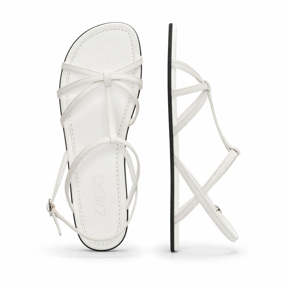 Introducing the QADA White Sandals from ZAQQ, made from high-quality nappa leather. These elegant barefoot sandals have thin leather straps and a smooth, flat sole. One sandal is seen from above, while the other shows off its sleek side profile with a small ankle strap buckle.