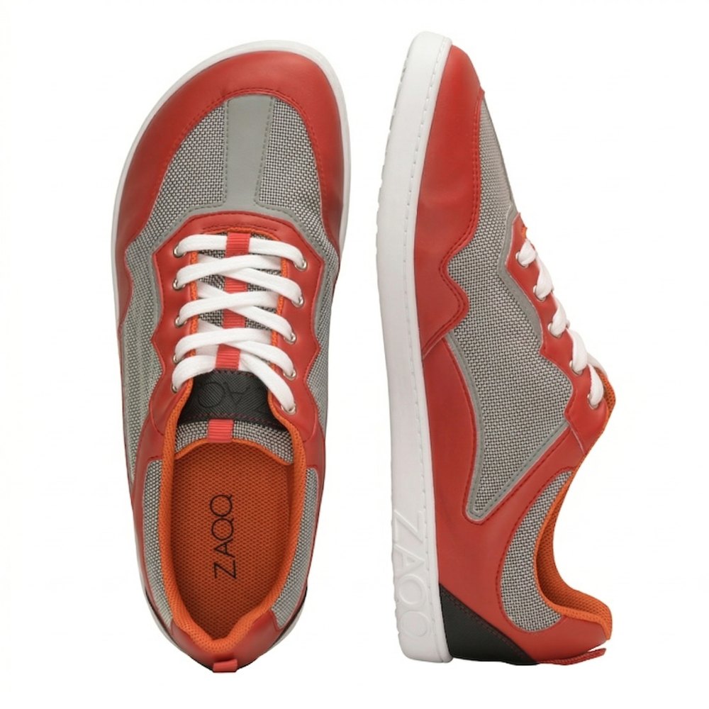 The QOPA Red Sneakers from ZAQQ impress with their eye-catching red-gray mesh design. A sneaker stands upright and lifts its white sole with the ZAQQlogo stands out while the other lies flat, showing off its orange interior and white laces. These barefoot shoes combine style with breathability for all-day comfort.