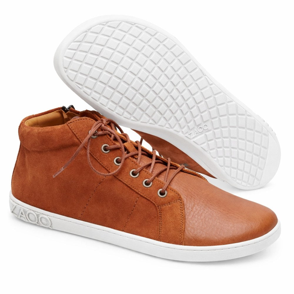 The QORE Mid Sierra from ZAQQ exudes elegance with its brown leather high-top design and beautifully contrasting white soles. One shoe stands upright while the other features a textured pattern on the sole that highlights the different types of leather used in its creation.